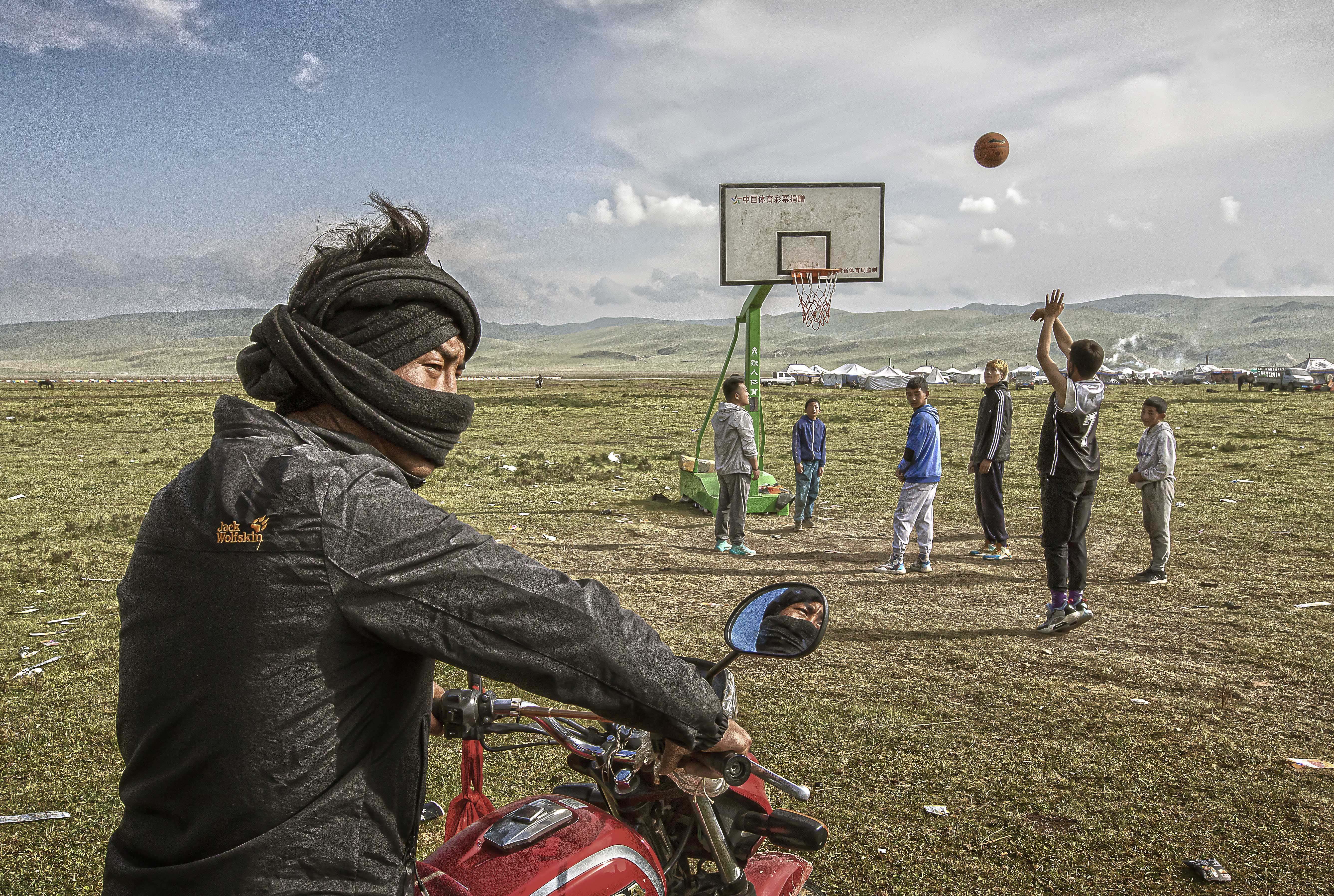 A man on a motorcycle in Tibet. Others play basketball in the background.