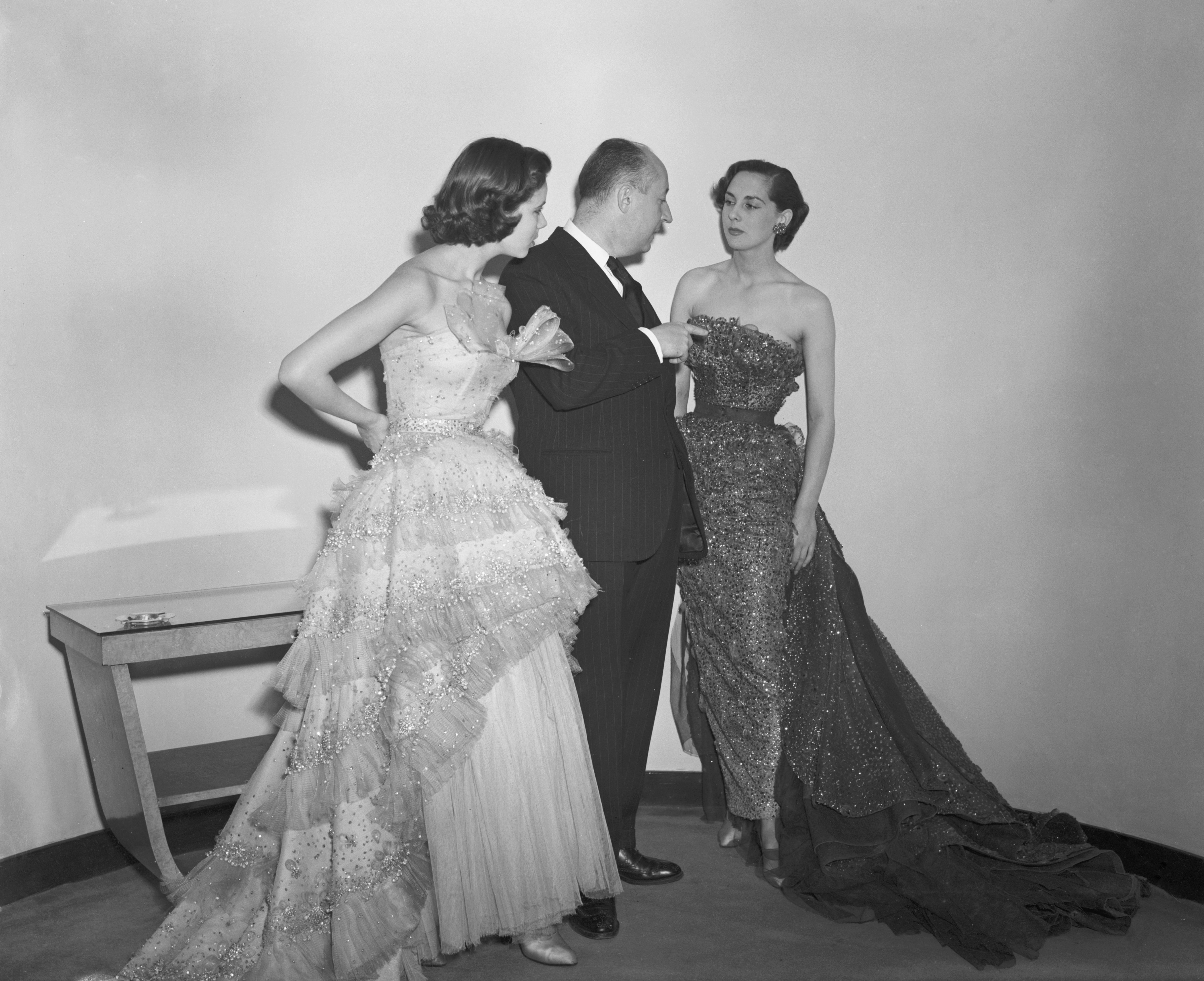 French fashion designer Christian Dior (1905 - 1957) with two of his models at a show of ninety new Dior creations at the Savoy hotel, London, 24th April 1950. Sylvie (left) models 'Mozart', a champagne tulle evening gown embroidered with diamante and straw. Russian model, Tania, models 'Debussy', a dark blue evening gown in tulle with sequins of the same colour. (Photo by Fred Ramage/Keystone/Hulton Archive/Getty Images)